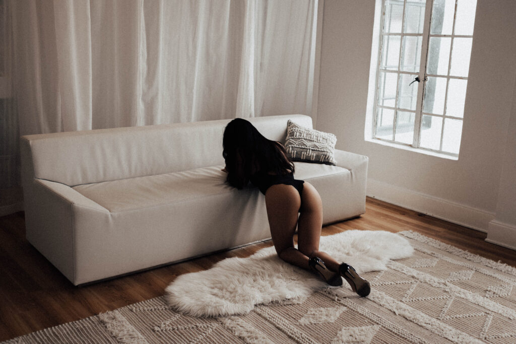 Woman in black lingerie posing confidently on floor beside white couch during Seattle boudoir photography session with natural light