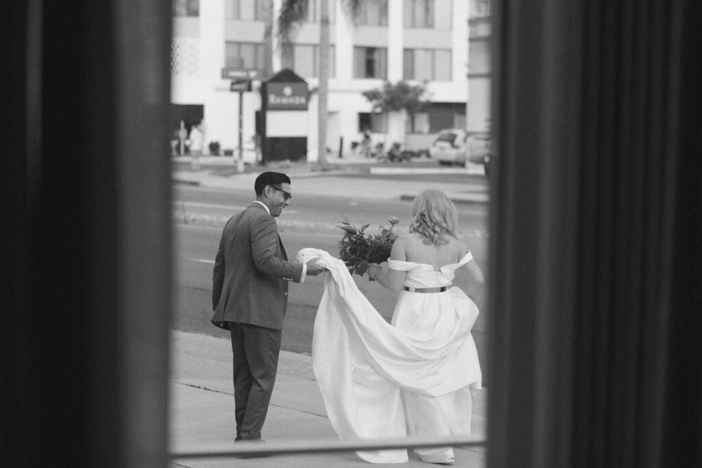 Candid black and white wedding photo, groom helping bride with her dress on city sidewalk, emotional wedding moment Seattle, documentary-style wedding photography Pacific Northwest, real wedding day moments