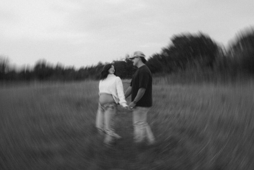 A maternity couple holding hands and smiling in a blurred motion photo taken in a rural field near Norman, Oklahoma. Captured by Rachael Marie Photography, this artistic maternity portrait highlights connection, movement, and love in the Oklahoma countryside.w