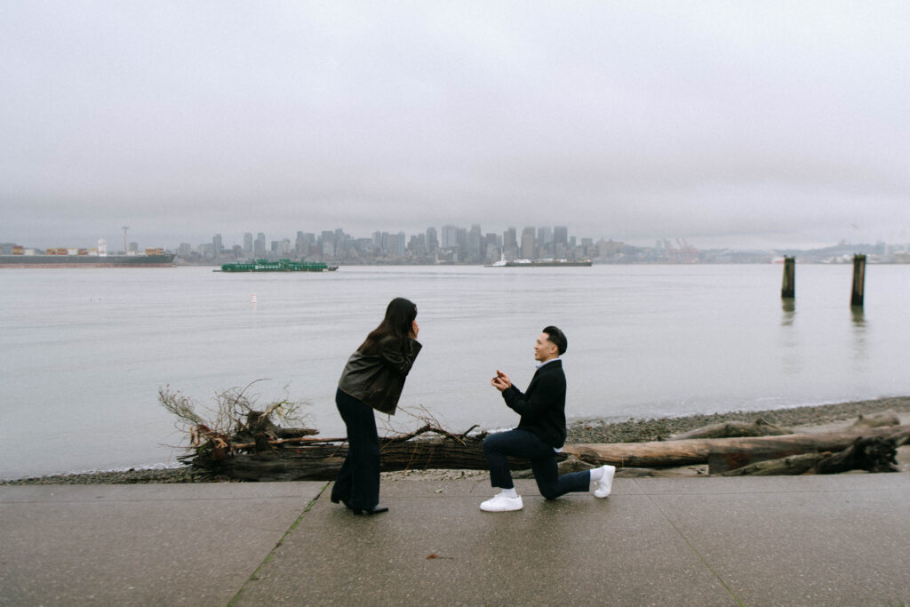 Man proposing during a surprise proposal at Alki Beach in West Seattle with the Seattle skyline in the background